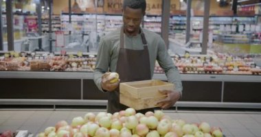 Afro-Amerikalı satıcı, elmaları tahta kutuya koyup süpermarkette teslimat için meyve hazırlıyor. Perakende ve manav konsepti.