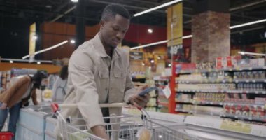 Afro-Amerikan adam akıllı telefon uygulaması kullanıyor alışveriş listesini kontrol ediyor ve süpermarketteki yiyecek ürünlerine bakıyor. Modern teknoloji ve satın alma kavramı.