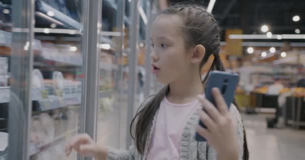 可爱的小女孩在网上用智能手机打电话 一边说一边指着超市里的食物 虚拟会议和互联网通信概念 — 图库视频影像