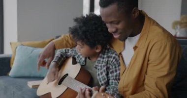 Neşeli Afro-Amerikalı çocuk, babasıyla gitar çalıyor. Evdeki kanepede oturup eğleniyor. Müzik aleti ve çocukluk konsepti.