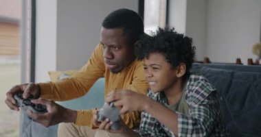 Afro-Amerikalı çocuk evde gülerek eğlenmek için babasıyla video oyunu oynuyor. Neşeli çocuk ve mutlu baba kavramı.