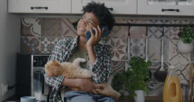Neşeli Afrikalı Amerikalı çocuk cep telefonuyla konuşuyor. Gülümsüyor. Mutfak masasında oturan kediciği tutuyor. Çocukluk ve iletişim kavramı.