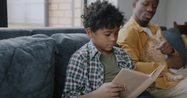 Feliz Niño Afroamericano Leyendo Libro Mientras Papá Acariciando Gato Sentado — Vídeo de stock