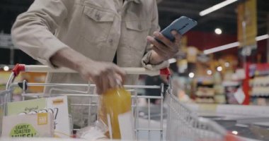 Süpermarkette erkek el alışverişine yakın çekim. Arabaya yiyecek koyar ve akıllı telefon kullanır. Modern teknoloji ve tüketim kavramı.