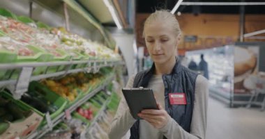 Envanter ve kalite kontrolü yapan kadın çalışan tablet kullanarak süpermarketteki raflarda ürünleri kontrol ediyor. Modern teknoloji ve perakende ticaret kavramı.