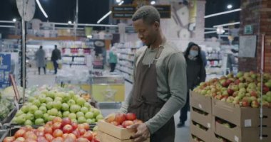 Afrika kökenli Amerikalı bir adamın önlüklü süpermarket çalışanı elinde elma kutusuyla rafa meyve koyması. Bakkal ve perakende sektörü kavramı.