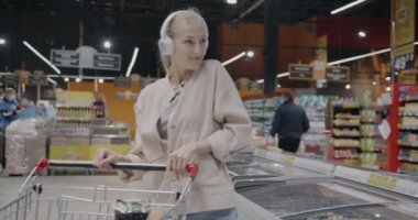 Süpermarkette dans eden neşeli genç bayan kulaklıkla müzik dinleyip yemekle eğleniyor. Modern yaşam tarzı ve alışveriş anlayışı.