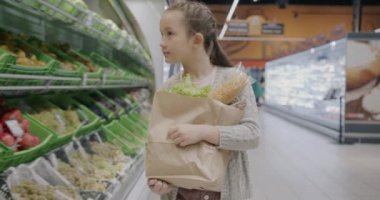 Süpermarkette elinde kese kağıdıyla yürüyen tatlı küçük kız tek başına meyve alışverişine bakıyor. Çocukluk ve tüketim kavramı.