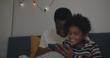 Baba ve oğul akıllı telefon kullanıyor ve gülüyorlar. Karanlıktaki yatak odasında oturmuş birlikte eğleniyorlar. Aile yaşam tarzı ve aygıt konsepti.