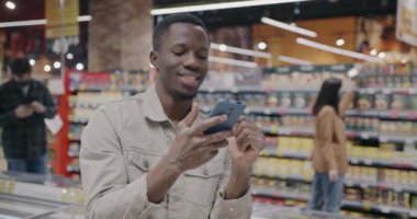Modern bir süpermarkette akıllı telefon mesajları kullanan Afro-Amerikalı bir adam. Modern iletişim teknolojisi ve alışveriş kavramı.