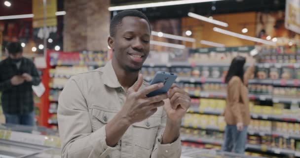 現代のスーパーマーケットの屋内でスマートフォンメッセージングを使用するアフリカ系アメリカ人男性 現代コミュニケーション技術とショッピングコンセプト — ストック動画