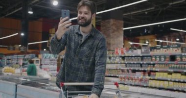 Neşeli adam süpermarketten alışveriş yapıyor ve akıllı telefon kullanarak internetten yemek tartışıyor. Uzak iletişim ve perakende kavramı.