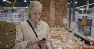 Ticari dokümanları okuyan ve markette yiyecekleri işaret eden ürünleri kontrol eden genç bir kadın. Perakende mağazası ve ticaret kavramı.