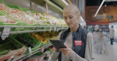 Tablet kullanan genç kadın süpermarket müdürünün portresi sonra raftaki yiyecekleri kontrol eden kameraya bakıyor. Modern teknoloji ve perakende ticaret kavramı.