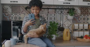 Yavru kedi tutan ve mutfak masasında akıllı telefon kullanan mutlu Afro-Amerikan çocuğun ağır çekimleri. Çocukluk ve alet kavramı.