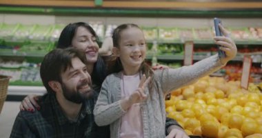 Kadın erkek ve küçük kız mutlu bir aile süpermarkette baş parmak hareketi yaparak selfie çekiyorlar ve eğleniyorlar. İnsanlar ve alışveriş konsepti.