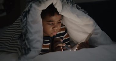 Küçük mutlu çocuk elinde fener tutarak kitap okuyor. Karanlık yatak odasındaki sıcak battaniyenin altında gülümsüyor. Çocukluk ve edebiyat kavramı.