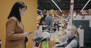 Yüz maskesi takan bir kadın, süpermarkette akıllı telefonuyla ödeme yapıyor. Kağıt torba alırken kasiyer alışverişi kabul ediyor. Alışveriş ve modern teknoloji konsepti.