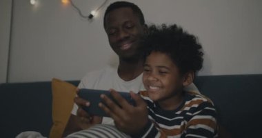 Akıllı telefonuyla online video oyunu oynayan şirin bir çocuk. Babasıyla yatakta oturmuş, yatak odasında eğleniyor. Oyun ve mutlu çocukluk konsepti.