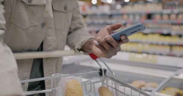 スマートフォンを使用して男性の手を閉じ スーパーマーケットで食品を購入しながら ショッピングトロリーで製品をチェックします 現代のガジェットと小売コンセプト — ストック動画