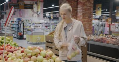 Elma satın alan genç bir kadın modern süpermarkette torbaya meyve koyuyor. Tüketim ve organik ürün satışı kavramı.