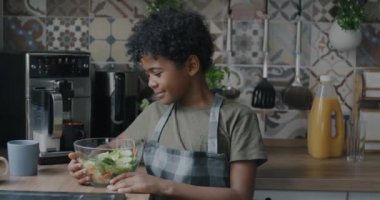Mutfakta kameraya gülümseyen bir kase salatayla önlük giymiş Afro-Amerikan çocuğun yavaş çekimde portresi. Çocukluk ve sağlıklı beslenme kavramı.
