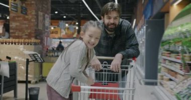 Çocuk eğlenirken küçük kız çocuğuyla alışveriş arabasını iten genç bir adamın ağır çekimde portresi. Aile ve mağaza müşteri konsepti.