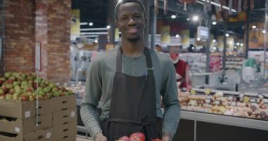 Süpermarkette elinde elma kutusuyla dikilen Afrikalı Amerikalı bir tezgahtarın yavaş çekimde portresi. Market ve meyve satma konsepti.