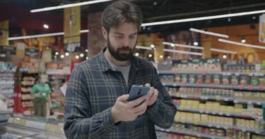 Süpermarkette duran ve ekranda akıllı telefon kullanan genç bir adamın portresi. Modern teknoloji ve perakende mağaza kavramı.