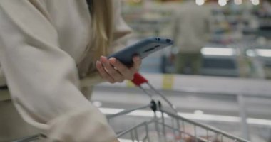 Akıllı telefon uygulamalarında alışveriş listesini kontrol eden ve süpermarketteki tramvaydaki yiyeceklere dokunan kadınların yakın çekimleri. Modern alet ve tüketim konsepti.