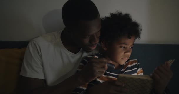 爱爸爸的小男孩晚上坐在床上 手里拿着手电筒 给他读一本有趣的书 童年和家庭生活方式概念 — 图库视频影像