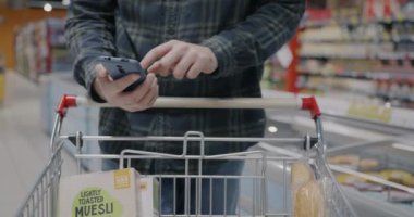 Erkek eli akıllı telefon kullanarak ve modern süpermarkette alışveriş arabasındaki ürünleri kontrol ederek. Teknoloji ve perakende kavramı.