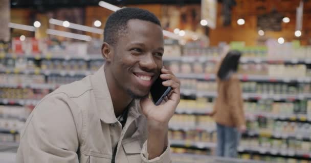 アフリカ系アメリカ人の男性は スーパーマーケットでの購入を議論している携帯電話で話す顧客を満足させました 携帯電話のコミュニケーションとショッピングのコンセプト — ストック動画