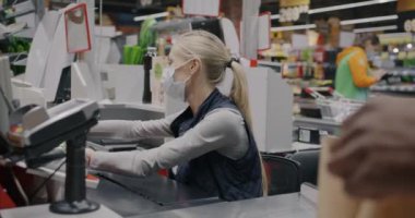 Yüz maskesi takan kadın kasiyer ürünleri tarıyor ve salgın sırasında gişede çalışan müşterilere yemek veriyor. Alışveriş ve perakende iş konsepti.