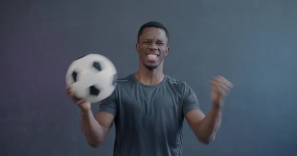 グレーの背景に喜びを表現する勝利を祝うサッカーを開催する興奮するアフリカ系アメリカ人の男性のゆっくりとした動きの肖像画 スポーツと感情の概念 — ストック動画