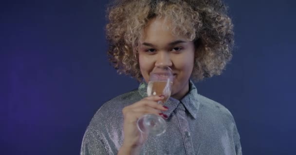 グラスからシャンパンを飲むアフリカ系アメリカ人の女性のゆっくりとした動きの肖像画は 青い背景のカメラを見ながら笑っている アルコールと感情コンセプト — ストック動画