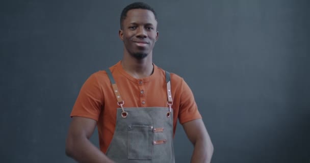 エプロンのアフリカ系アメリカ人のスローモーション肖像画と灰色の背景に立っている笑顔 小規模ビジネスとキャレッティングのコンセプト — ストック動画
