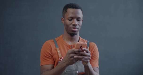 エプロンのアフリカ系アメリカ人男性は グレー色の背景にモバイルアプリケーションを使用してスマートフォンテキストを使用しています モダンなテクノロジーと起業家のスタートアップコンセプト — ストック動画