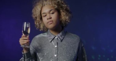 Mavi arka planda köpüklü şampanya bardağıyla dans eden Afro-Amerikan bayanın yavaş çekim portresi. Alkolik içki ve parti konsepti.