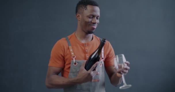 アフリカ系アメリカ人のウェイターがグラスにワインを注ぎ 灰色の背景でカメラを見る アルコール配送と小規模ビジネスコンセプト — ストック動画