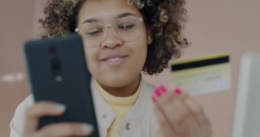 Akıllı telefonlu mutlu Afro-Amerikan bayan alışverişi çevrimiçi mağazadan alıyor evde gülümseyen kredi kartıyla ödüyor. Gadget ve tüketim kavramı.
