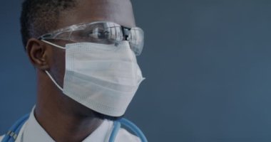 Yüz maskesi takmış Afrikalı Amerikalı bir doktorun yakın çekim yavaş çekim portresi ve gri arka planda kameraya yansıyan gözlükler. Sağlık ve tıp mesleği kavramı.