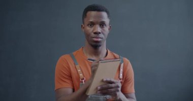 Önlüklü Afro-Amerikan bir adamın portresi. Defterde yazıyor ve kameraya bakıyor. Sonra da gri arka plan üzerine kağıt veriyor.
