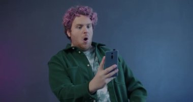 Mobil kameraya poz veren yaratıcı adamın yavaş çekim portresi. Akıllı telefonlu selfie çekiyor. Mor arka planda komik suratlar yapıyor.