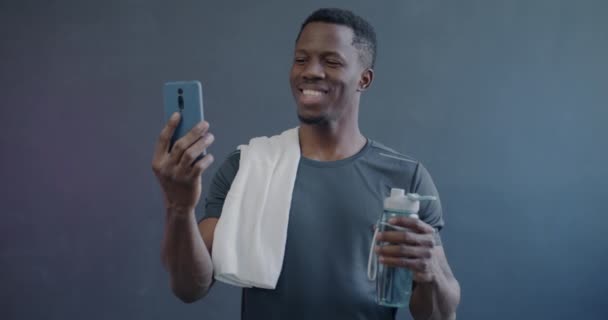 アフリカ系アメリカ人のスポーツマンがオンラインビデオ通話と紫色の背景のボトルからの飲料水を作る携帯電話で話す — ストック動画