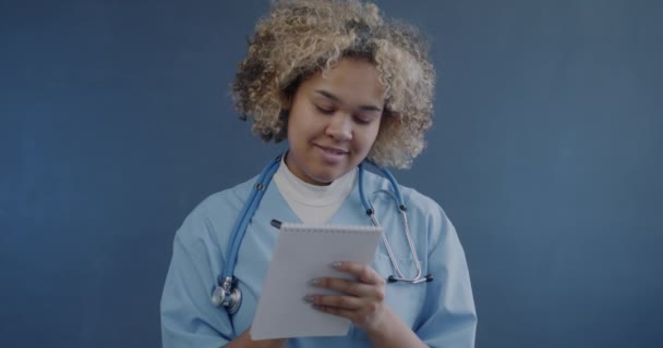 非洲裔美籍女医生的画像 她穿着制服 在笔记本上写着处方药 在蓝色背景下给相机配纸 — 图库视频影像