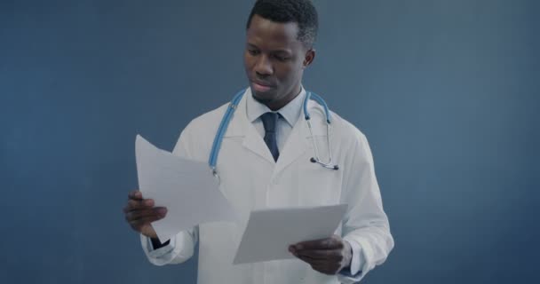 白いガウンのアフリカ系アメリカ人医師の肖像画は 灰色の背景に紙を保持する医学記録を読んでいます ヘルスケアとプロの職業コンセプト — ストック動画