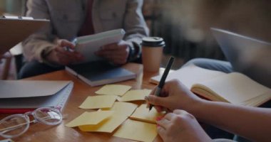 Masada çalışan bir grup insan başlangıç projesiyle meşgulken, kadın el yazısının yapışkan notlara yakın plan çekimleri. Yaratıcı iş ve insanların kavramı.