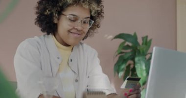 Afro-Amerikalı iş kadını dizüstü bilgisayarla yazı yazıyor ve kredi kartıyla gülümsüyor ve ofisteki internet finansmanından keyif alıyor. İş kadını ve bankacılık kavramı.