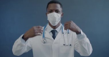 Beyaz önlüklü Afrikalı Amerikalı doktorun yüz maskesi çıkartıp gri arka plandaki kameraya baktığı yavaş çekim portresi. Tıp ve tekdüze kavram.